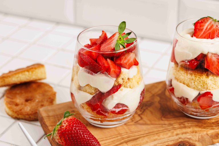 Two glass cups filled with layers of sliced strawberries, sponge cake, and creamy whipped topping create a delightful strawberry mascarpone trifle. A fresh strawberry and a round piece of cake rest nearby on a white tiled surface.