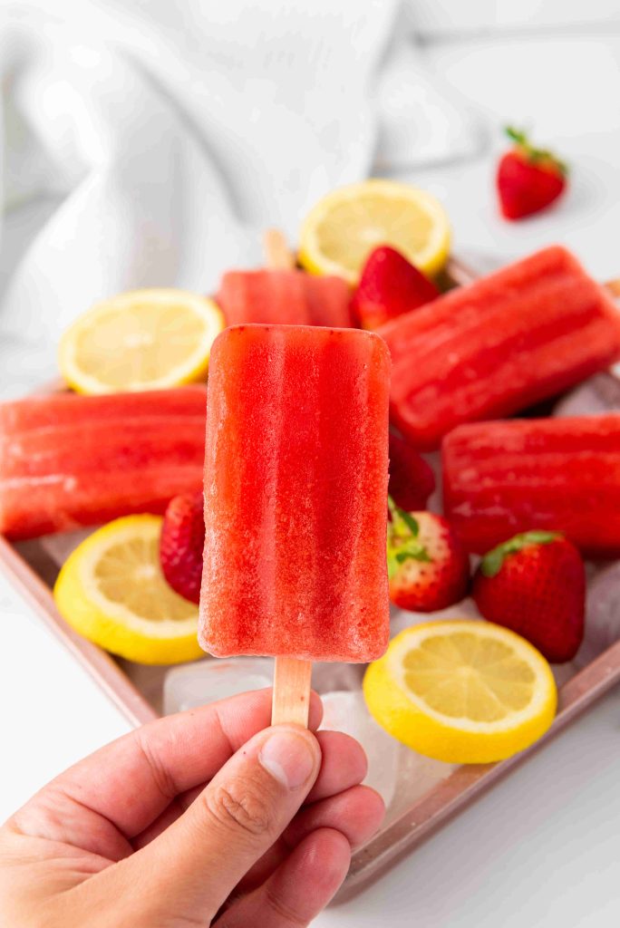 A hand holds a bright red popsicle in front of a tray filled with more Easy Strawberry Popsicles Recipe treats, lemon slices, and whole strawberries, all arranged on ice.