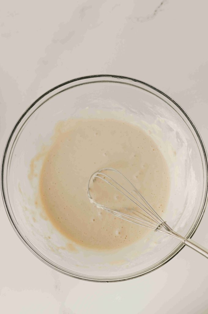A glass mixing bowl filled with pancake batter sits on a light countertop. A metal whisk, reminiscent of one used in an Old-Fashioned Strawberry Cobbler Recipe, is resting in the bowl, partially covered in batter.