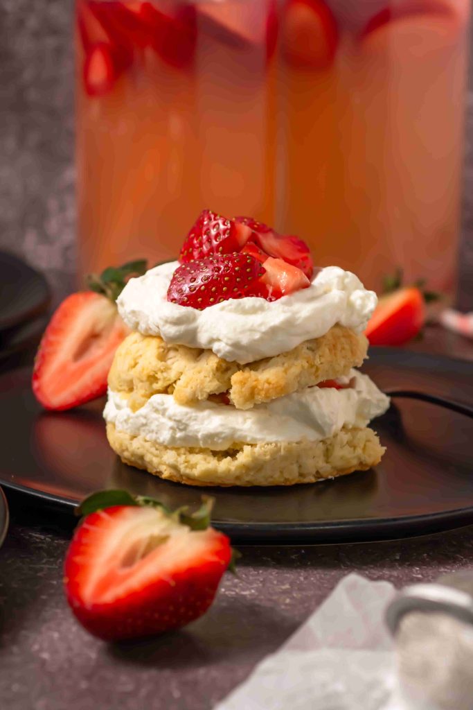 An Old Fashioned Strawberry Shortcake Recipe features a shortcake topped with whipped cream and sliced strawberries on a black plate, with halved berries and a pitcher of pink drink in the background.