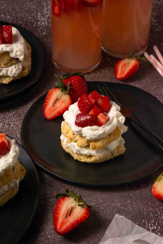 A black plate with an Old Fashioned Strawberry Shortcake Recipe, topped with whipped cream and fresh strawberries, surrounded by sliced berries, a fork, and glasses of pink drink on a dark textured surface.
