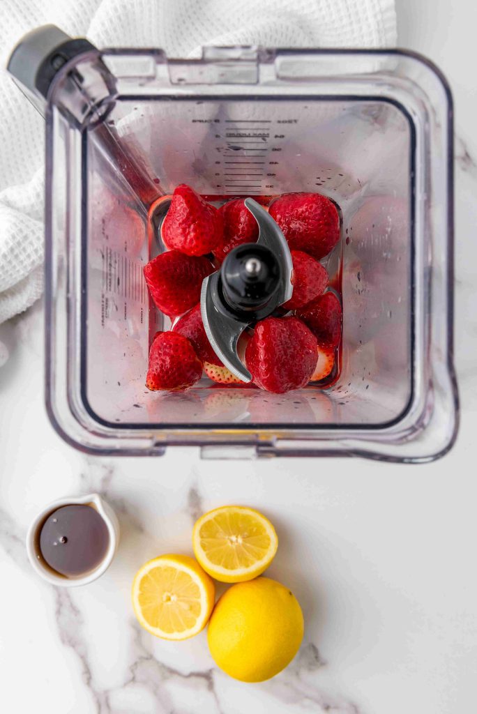 Overhead view of a blender with strawberries and liquid, ready for an Easy Strawberry Popsicles Recipe. On the white surface, a halved lemon, whole lemon, and small cup of syrup are neatly arranged—perfect for making Strawberry Lemonade Ice Pops.