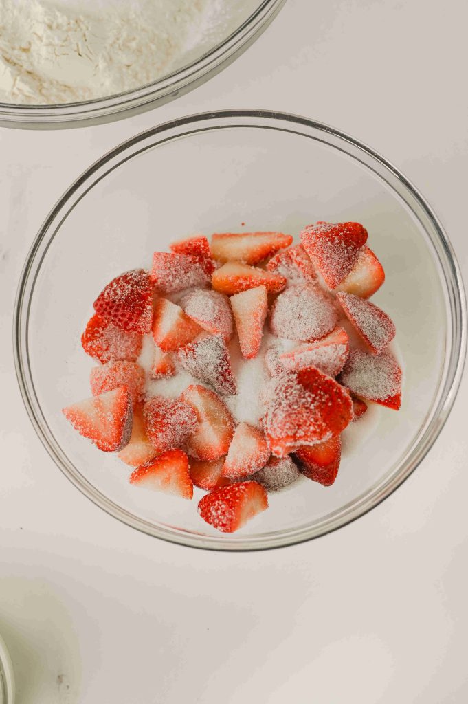 A glass bowl filled with sliced strawberries coated in granulated sugar, seen from above—perfect for starting an Old-Fashioned Strawberry Cobbler Recipe.