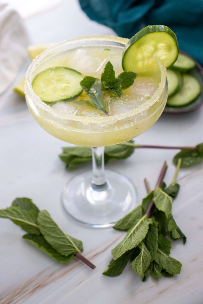 A Cucumber Margarita Mocktail in a margarita glass, garnished with cucumber slices and fresh mint, sits on a white surface ringed with mint sprigs. The rim of the glass is coated with sugar or salt for an extra refreshing touch.