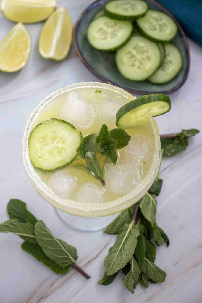 A glass of iced Cucumber Lime Mocktail garnished with cucumber slices and mint, surrounded by fresh mint sprigs, lime wedges, and a plate of cucumber slices on a light surface.