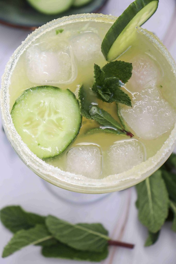 A close-up of a refreshing Cucumber Lime Mocktail with ice cubes, cucumber slices, fresh mint leaves, and a salted rim, viewed from above. Some mint leaves are scattered around the glass.