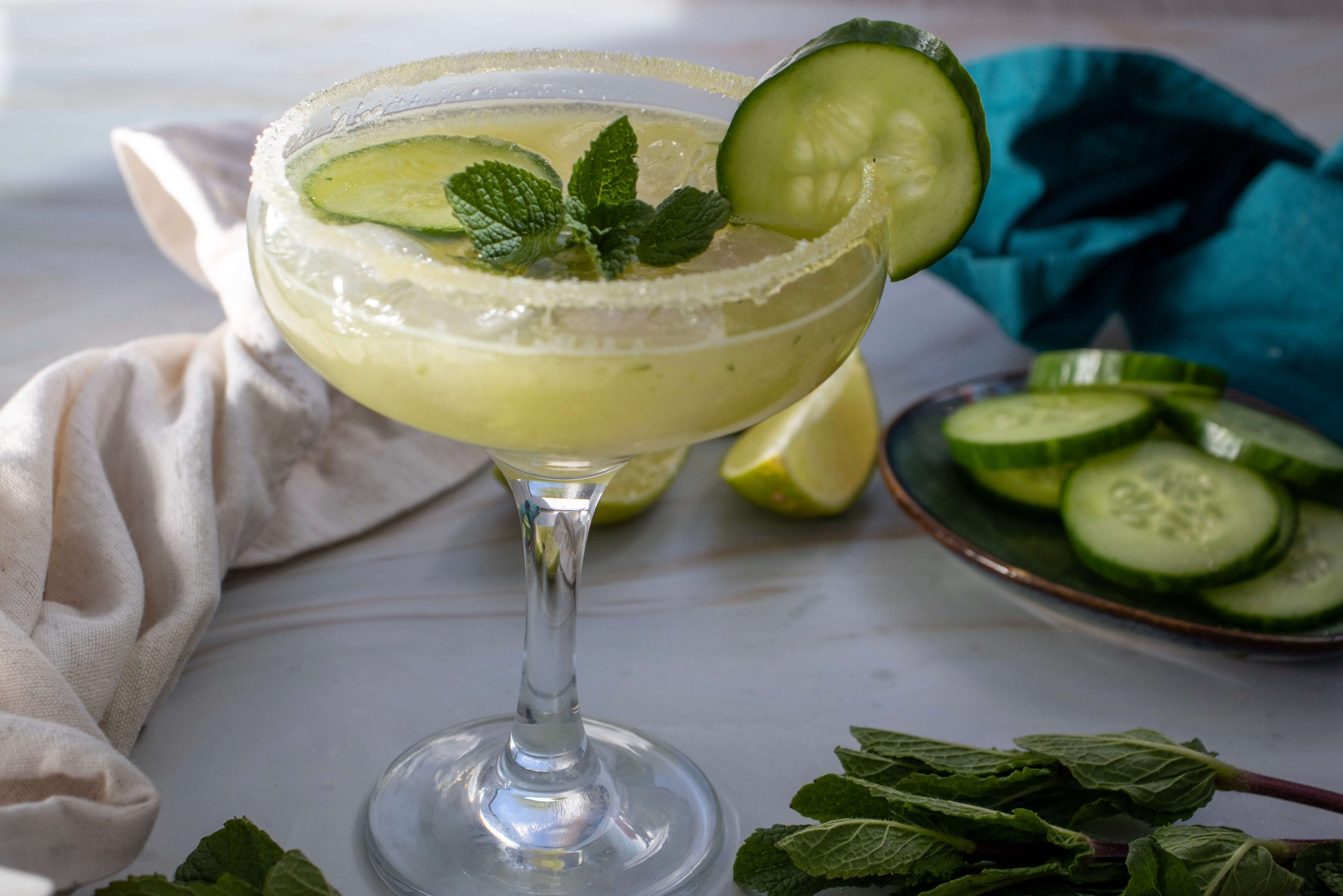 A margarita glass filled with a light green Cucumber Lime Mocktail, garnished with cucumber slices and fresh mint. A sugared rim, a plate of cucumbers, mint leaves, a lime wedge, and a blue cloth are visible in the background.