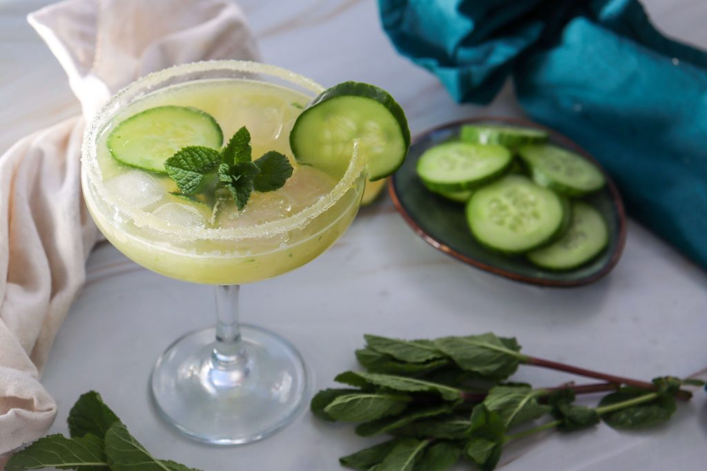 A margarita glass with a light green Cucumber Lime Mocktail, garnished with cucumber slices and fresh mint, sits on a white surface next to a plate of cucumber slices and sprigs of mint. A beige cloth and teal fabric are nearby.