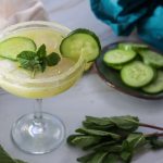 A margarita glass with a light green Cucumber Lime Mocktail, garnished with cucumber slices and fresh mint, sits on a white surface next to a plate of cucumber slices and sprigs of mint. A beige cloth and teal fabric are nearby.