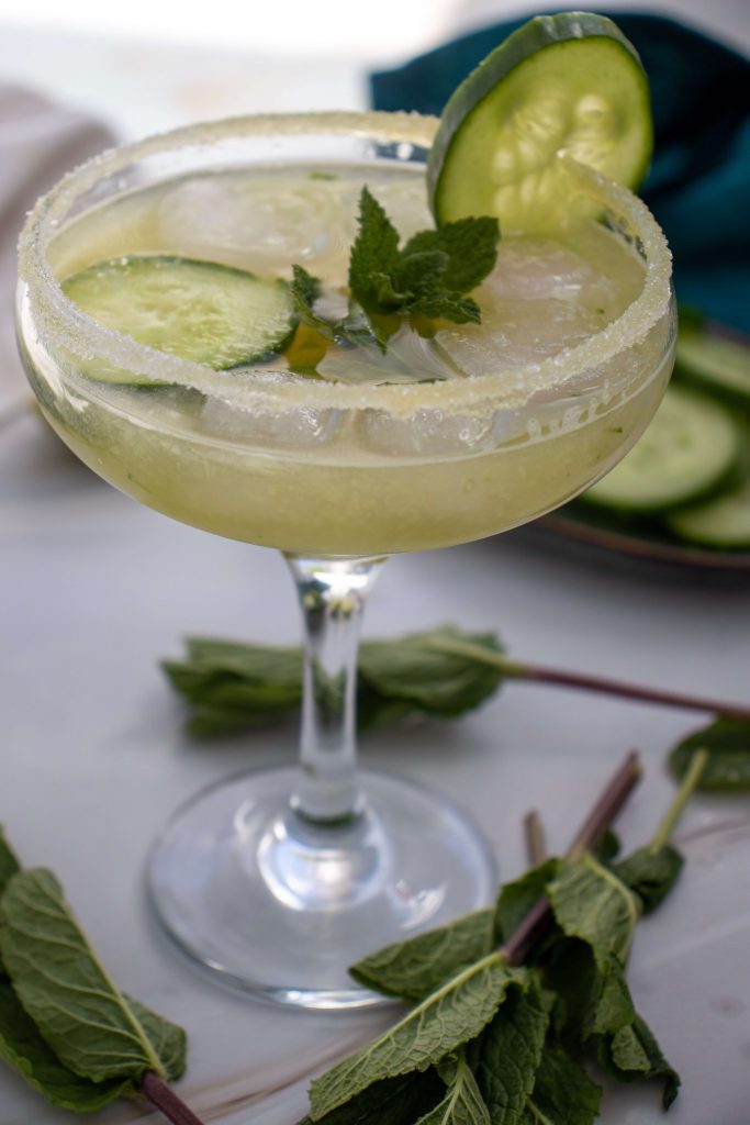 A Cucumber Margarita Mocktail in a coupe glass with a sugared rim, garnished with cucumber slices and fresh mint, sits on a white surface with mint sprigs nearby.