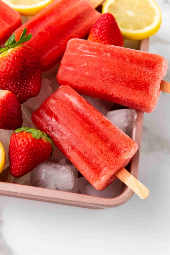 Bright red Strawberry Lemonade Ice Pops on ice in a pink tray, surrounded by fresh strawberries and lemon slices—an easy strawberry popsicles recipe perfect for summer refreshment.