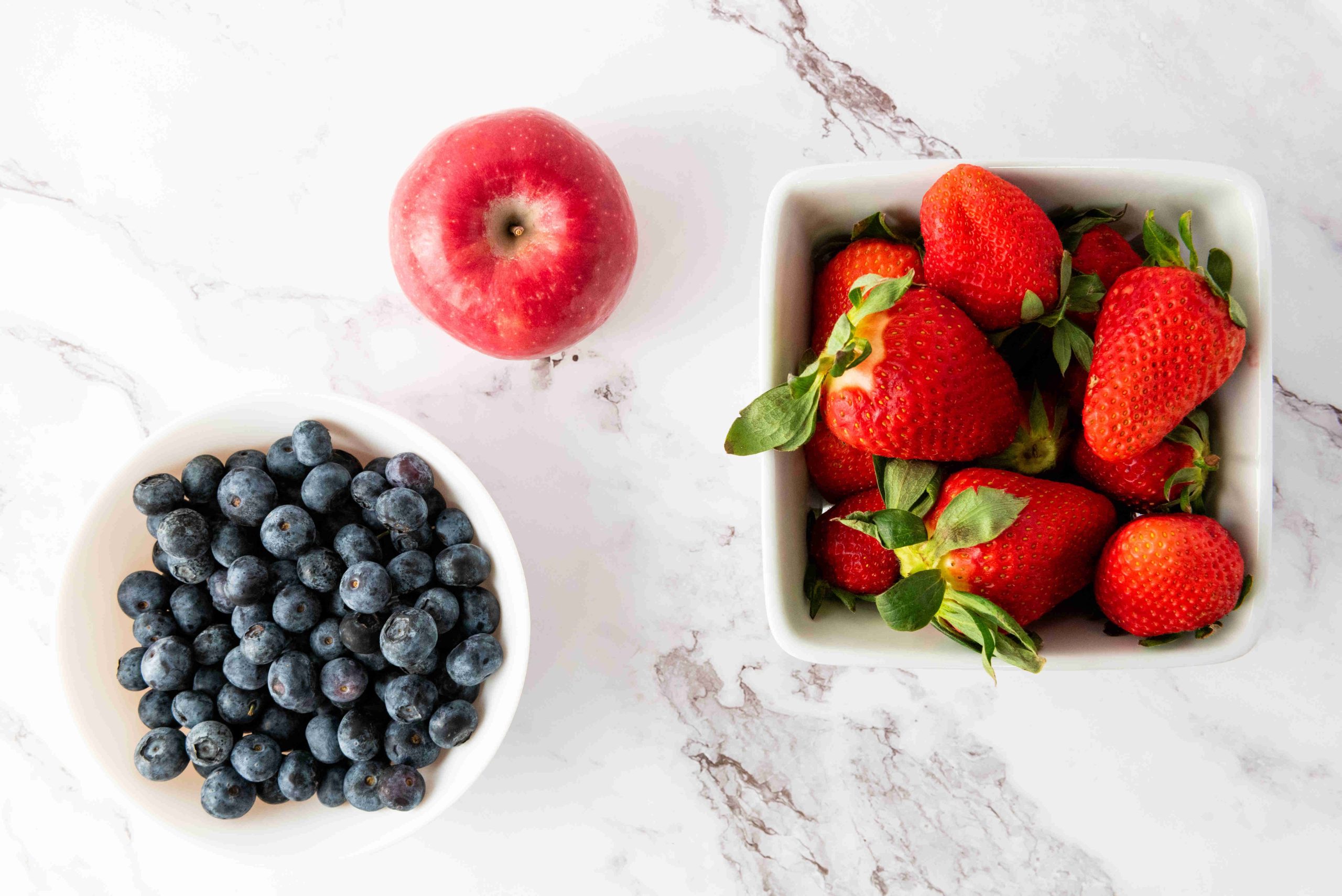 A bowl of fresh strawberries, a bowl of blueberries, and a red apple are arranged on a white marble surface, creating a patriotic fruit salad perfect for any occasion.