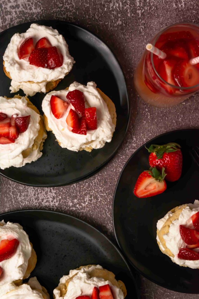 Top view of plates with an Old Fashioned Strawberry Shortcake Recipe, topped with whipped cream and sliced strawberries, next to a glass of strawberry drink with a straw, on a textured brown surface.