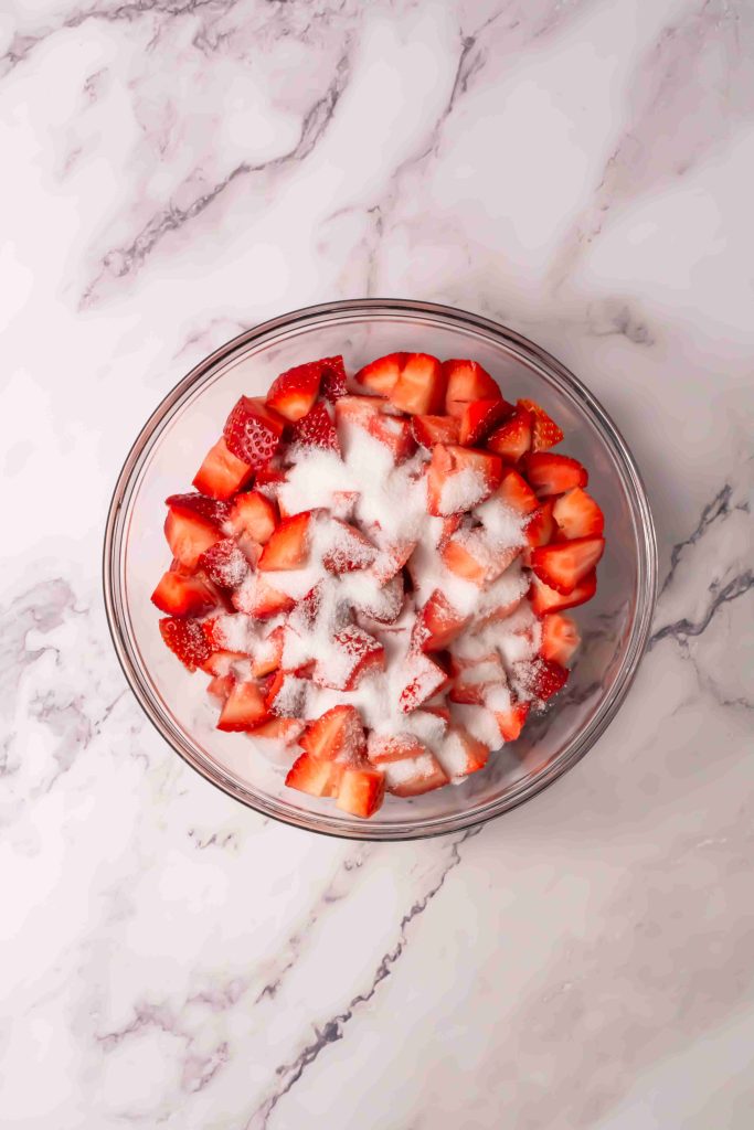 A glass bowl filled with chopped strawberries sprinkled with granulated sugar, just like in an Old Fashioned Strawberry Shortcake Recipe, placed on a white marble surface.