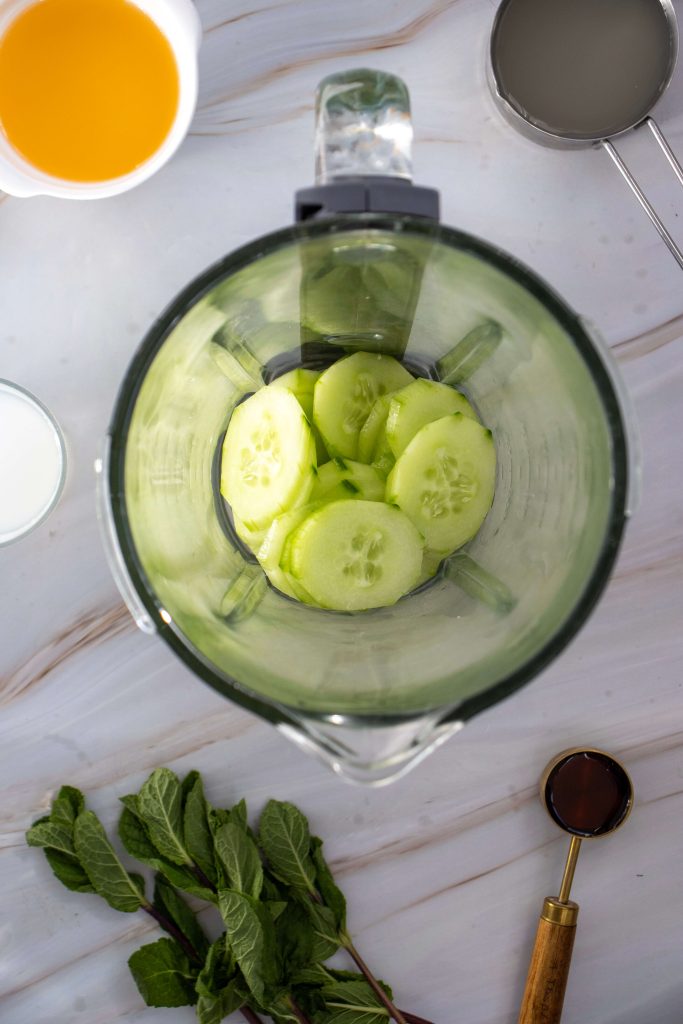 A top-down view of a blender with cucumber slices, perfect for making a refreshing Cucumber Margarita Mocktail, surrounded by measuring cups with liquids, fresh mint, and a tablespoon on a marble surface.
