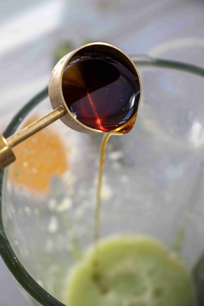 A metal measuring spoon pours dark liquid, likely soy sauce, into a glass bowl containing a light green mixture for a refreshing Cucumber Margarita Mocktail.