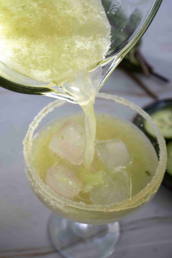 A close-up of a Cucumber Lime Mocktail being poured from a glass pitcher into a sugar-rimmed glass filled with ice cubes, with fresh cucumber slices visible in the background.