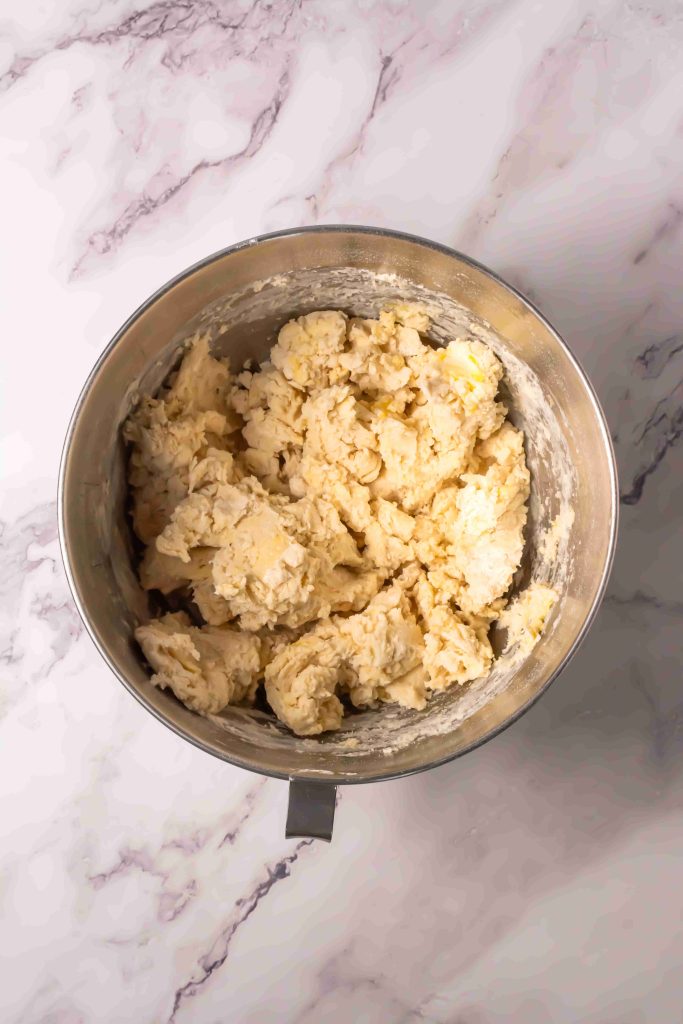 A metal mixing bowl filled with lumpy, partially mixed biscuit dough for an Old Fashioned Strawberry Shortcake Recipe sits on a white and gray marble countertop.