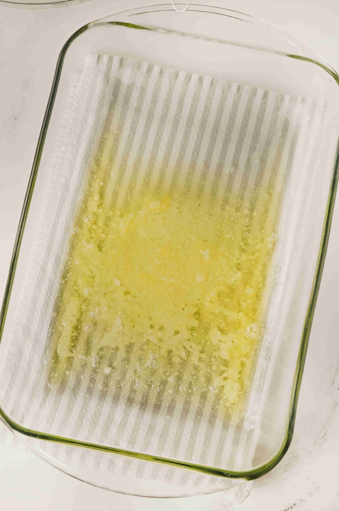 A glass baking dish lined with parchment paper and spread with melted butter, ready for the next step in this Old-Fashioned Strawberry Cobbler Recipe, viewed from above on a white surface.