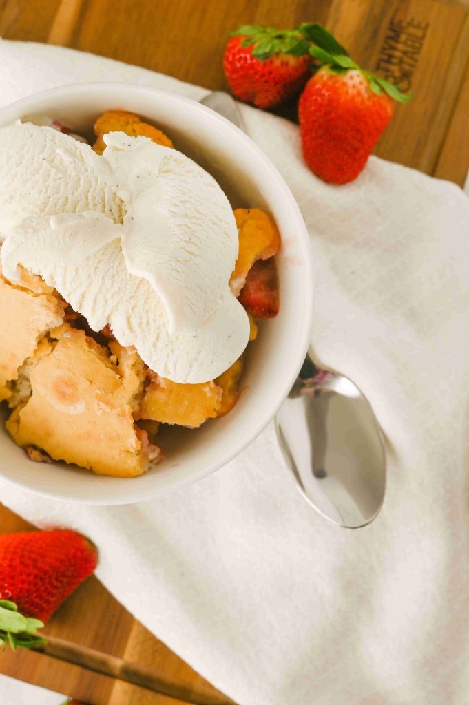 A bowl of Old-Fashioned Strawberry Cobbler Recipe topped with vanilla ice cream sits on a white cloth, with a spoon beside it and fresh strawberries nearby.