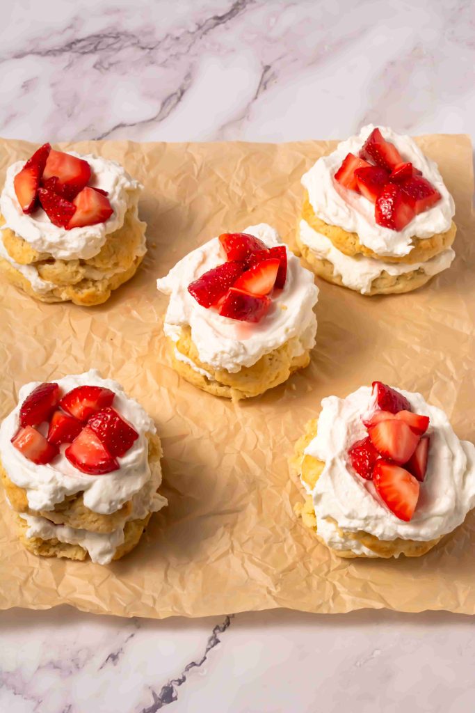 Five strawberry shortcakes, made from an old fashioned strawberry shortcake recipe, are topped with whipped cream and fresh strawberry pieces, arranged on a piece of parchment paper over a marble surface.