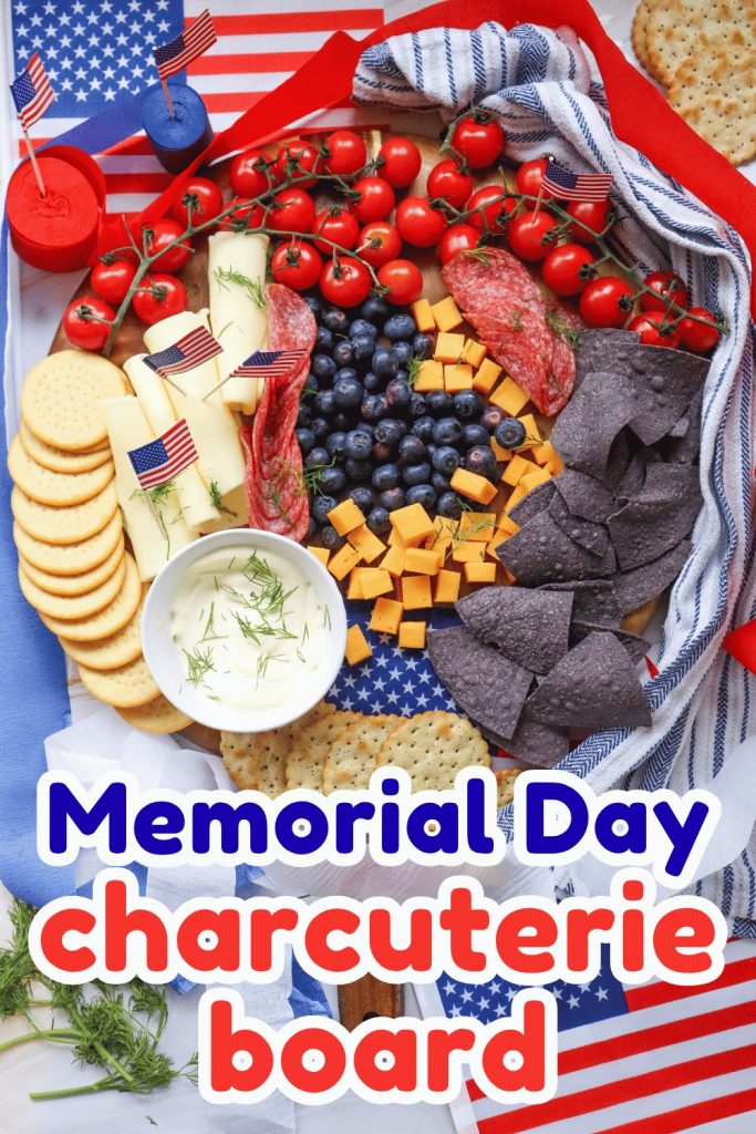 A festive Memorial Day Charcuterie Board decorated with mini American flags, featuring crackers, cheese cubes, tomatoes, blueberries, salami, blue corn chips, dip, and strawberries in a patriotic red, white, and blue theme.