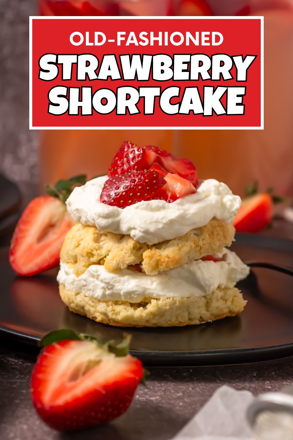 A biscuit-style strawberry shortcake topped with whipped cream and sliced strawberries sits on a black plate, surrounded by whole and halved berries. A bold sign above reads Old Fashioned Strawberry Shortcake Recipe.
