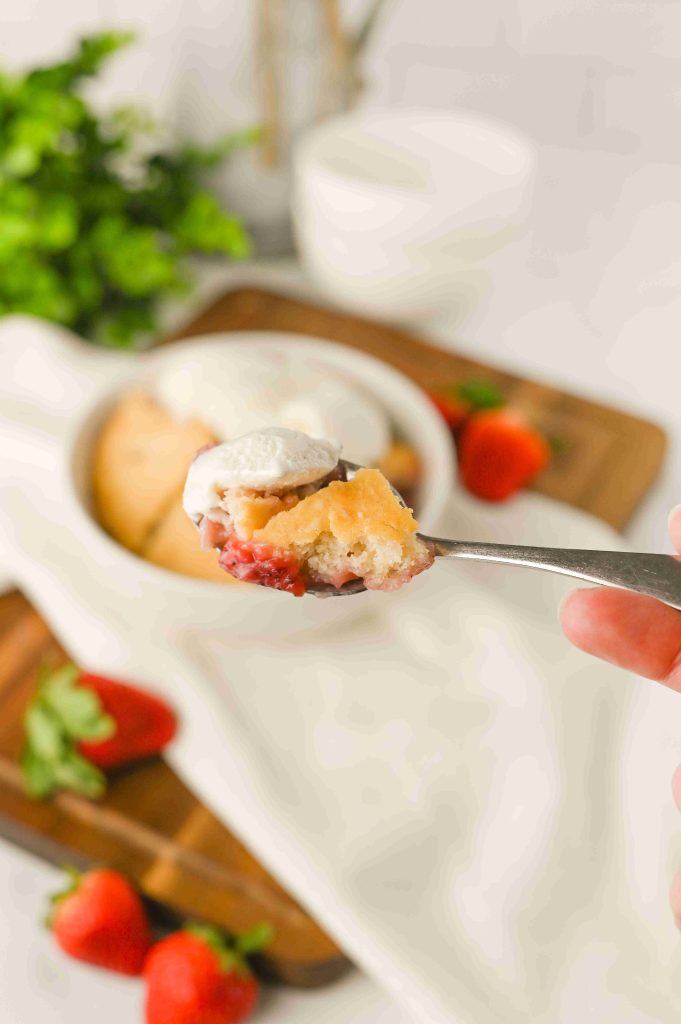 A close-up of a spoonful of strawberry shortcake topped with whipped cream, reminiscent of an Old-Fashioned Strawberry Cobbler Recipe, with a bowl of shortcake and fresh strawberries blurred in the background.