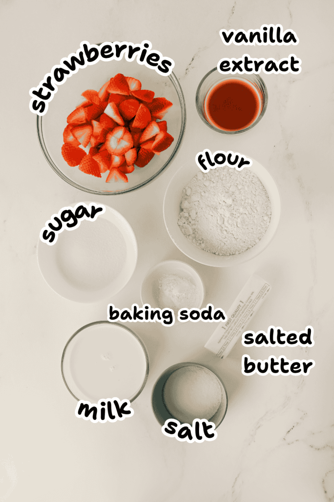 A top-down view of labeled baking ingredients in bowls on a white surface for an Old-Fashioned Strawberry Cobbler Recipe: sliced strawberries, vanilla extract, flour, sugar, baking soda, milk, salt, and salted butter.