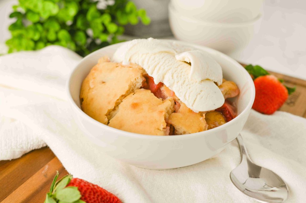 A bowl of old-fashioned strawberry cobbler topped with vanilla ice cream sits on a white cloth, with fresh strawberries and a spoon nearby. Greenery is blurred in the background, highlighting this classic dessert recipe.