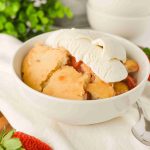 A bowl of old-fashioned strawberry cobbler topped with vanilla ice cream sits on a white cloth, with fresh strawberries and a spoon nearby. Greenery is blurred in the background, highlighting this classic dessert recipe.