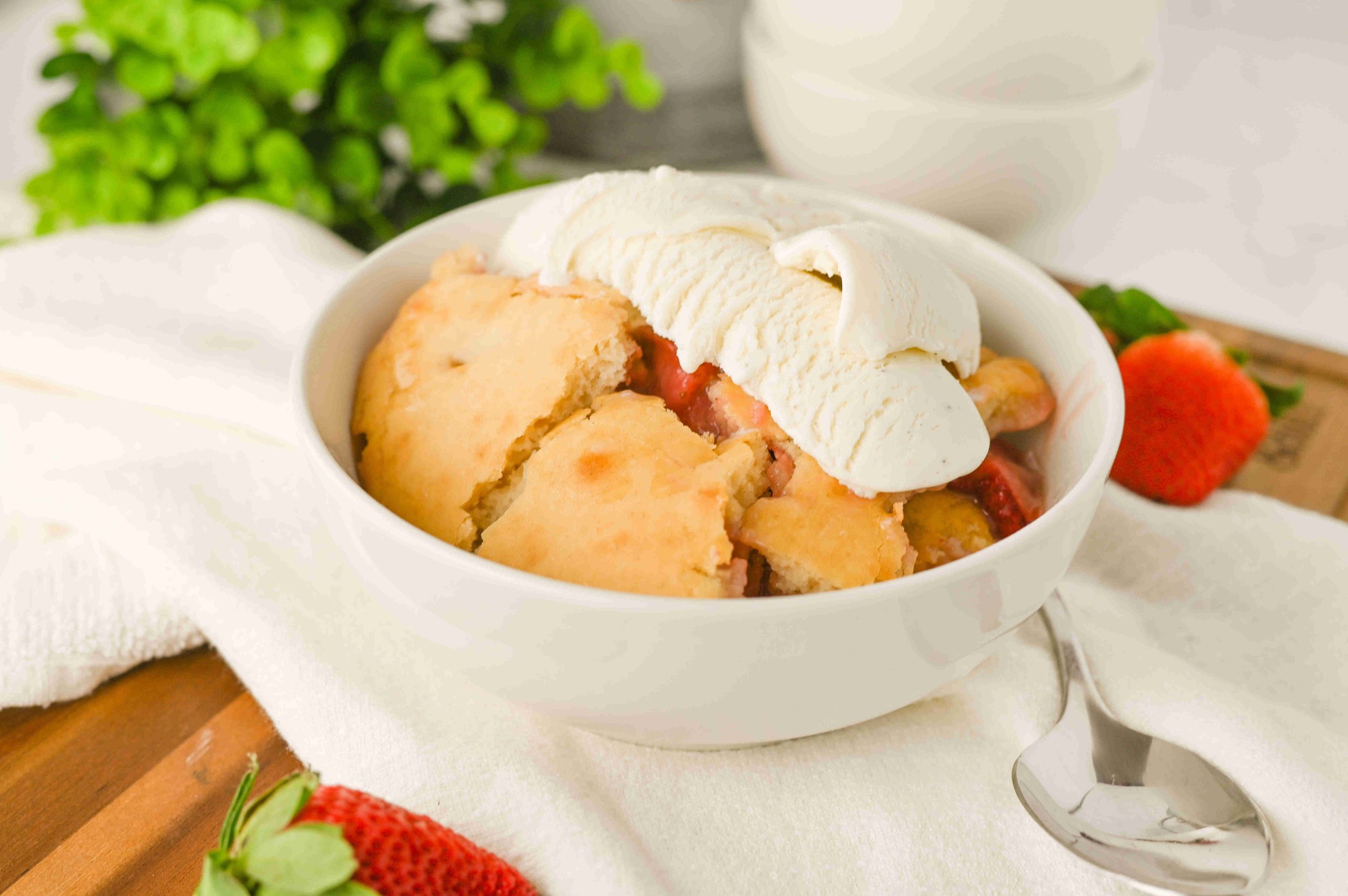 A bowl of old-fashioned strawberry cobbler topped with vanilla ice cream sits on a white cloth, with fresh strawberries and a spoon nearby. Greenery is blurred in the background, highlighting this classic dessert recipe.
