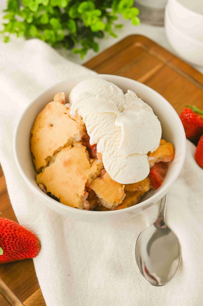 A bowl of Old-Fashioned Strawberry Cobbler Recipe topped with three scoops of vanilla ice cream sits on a white napkin next to a spoon, with fresh strawberries and a green plant in the background.