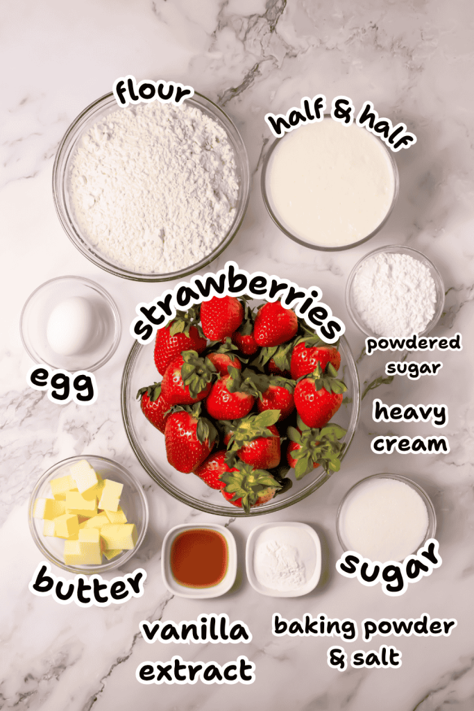 A flat lay of baking ingredients on a marble surface, perfect for an Old Fashioned Strawberry Shortcake Recipe—featuring flour, half & half, powdered sugar, heavy cream, sugar, cubed butter, an egg, vanilla extract, baking powder & salt, and fresh strawberries.