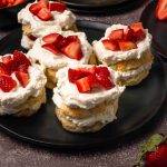 Four strawberry shortcakes, made from an Old Fashioned Strawberry Shortcake Recipe, are topped with whipped cream and fresh strawberry pieces on a black plate, with extra strawberries and a fork nearby on a dark surface.