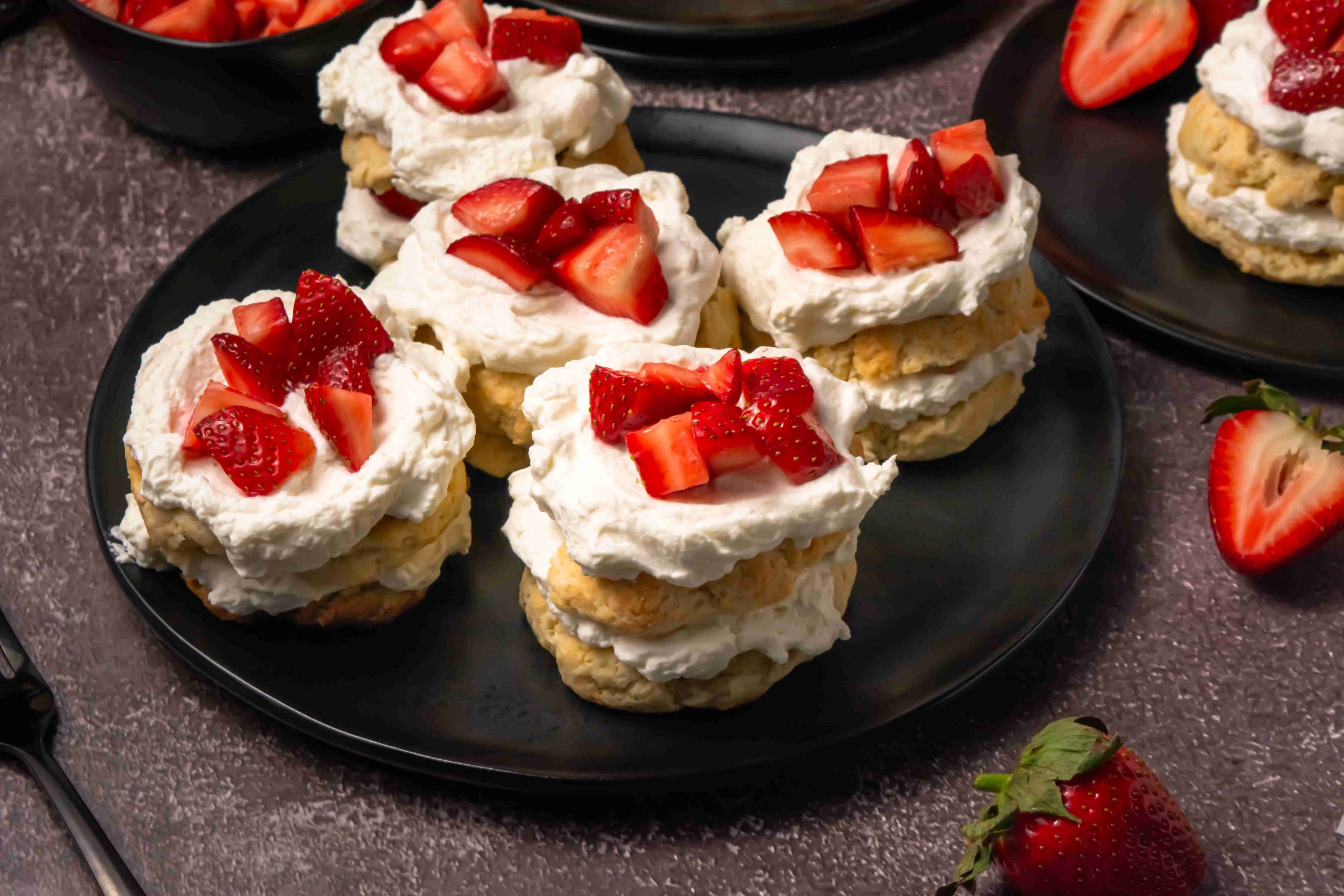 Four strawberry shortcakes, made from an Old Fashioned Strawberry Shortcake Recipe, are topped with whipped cream and fresh strawberry pieces on a black plate, with extra strawberries and a fork nearby on a dark surface.