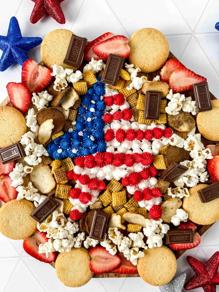 A patriotic snack board arranged with cookies, popcorn, strawberries, chocolate bars, and cereal, featuring a central star decorated with red, white, and blue icing to resemble the American flag.