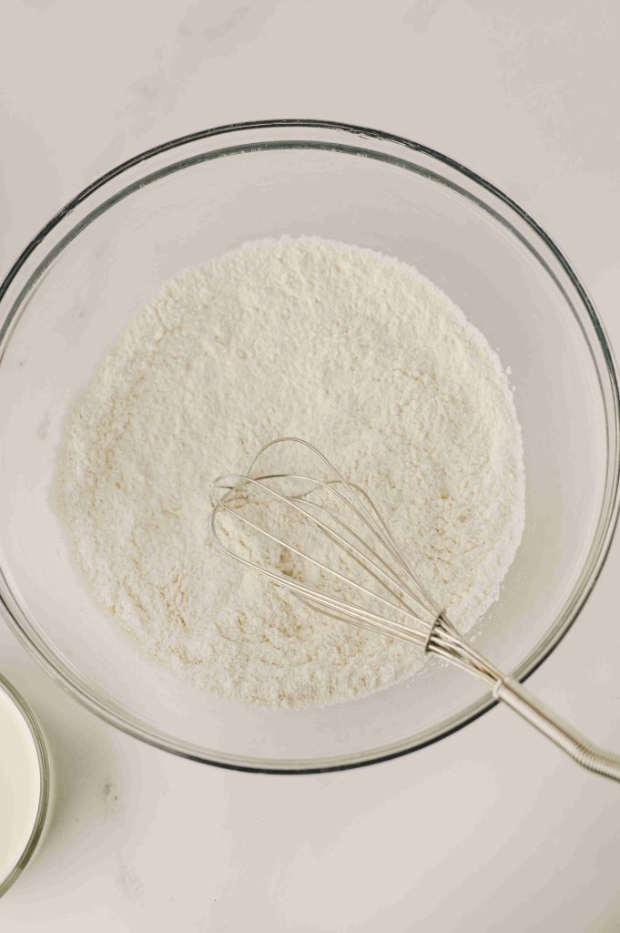 A whisk rests in a glass bowl filled with flour mixture on a light surface, ready for the Old-Fashioned Strawberry Cobbler Recipe. A small glass of milk sits nearby, hinting at the delicious baking to come.