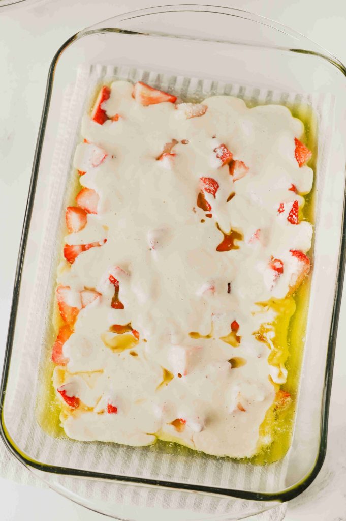 A glass baking dish filled with sliced strawberries and a creamy white batter for an Old-Fashioned Strawberry Cobbler Recipe sits on a white surface, the uncooked mixture slightly marbled with melted butter around the edges.