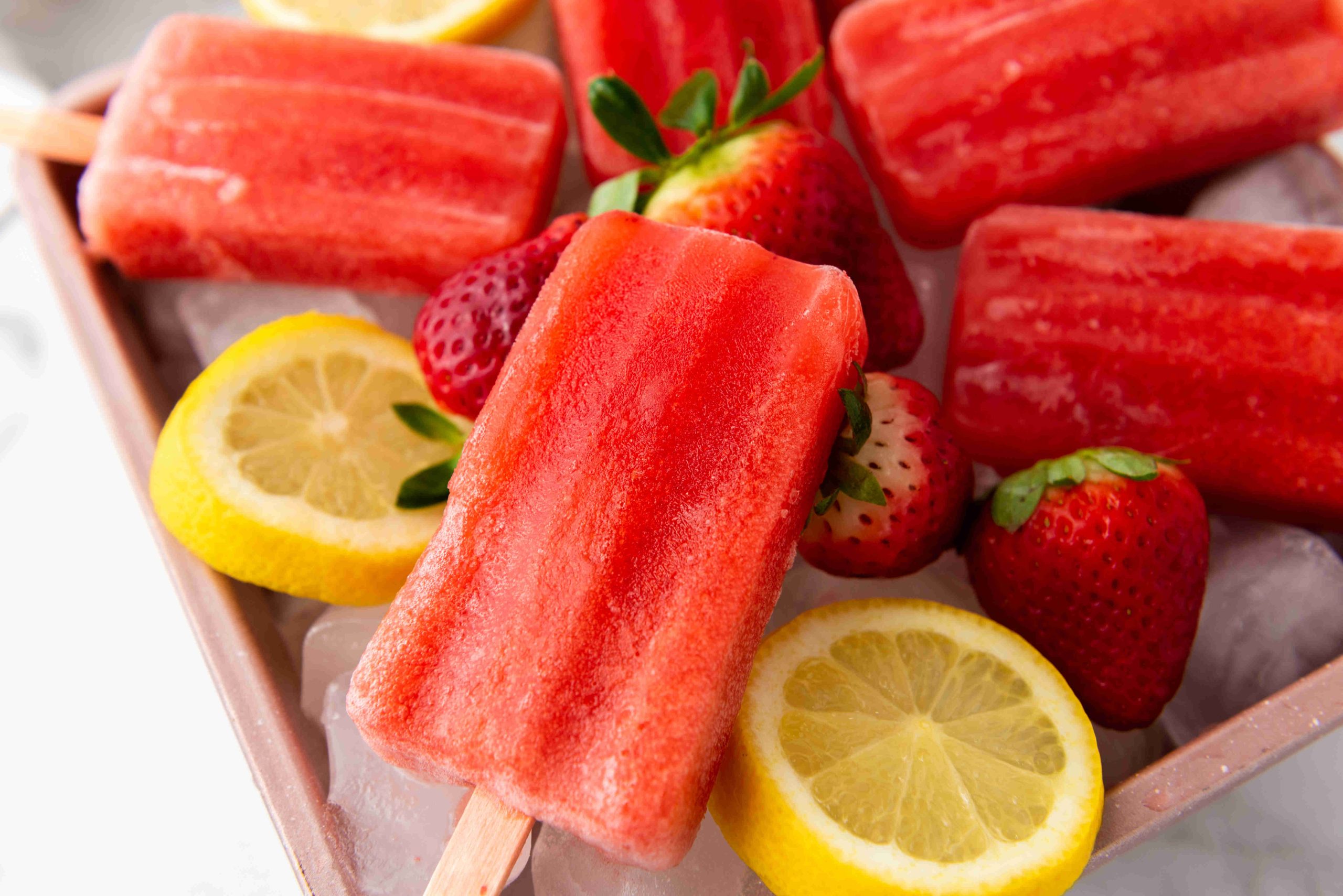 Strawberry popsicles arranged on ice with fresh strawberries and lemon slices around them. These vibrant treats are the perfect result of an Easy Strawberry Popsicles Recipe, making summer snacking simple and refreshing.