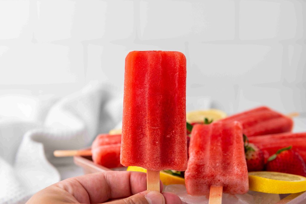 A hand holds a red popsicle in the foreground, with more Strawberry Lemonade Ice Pops, sliced lemons, and strawberries on a plate in the background against a white tiled wall.
