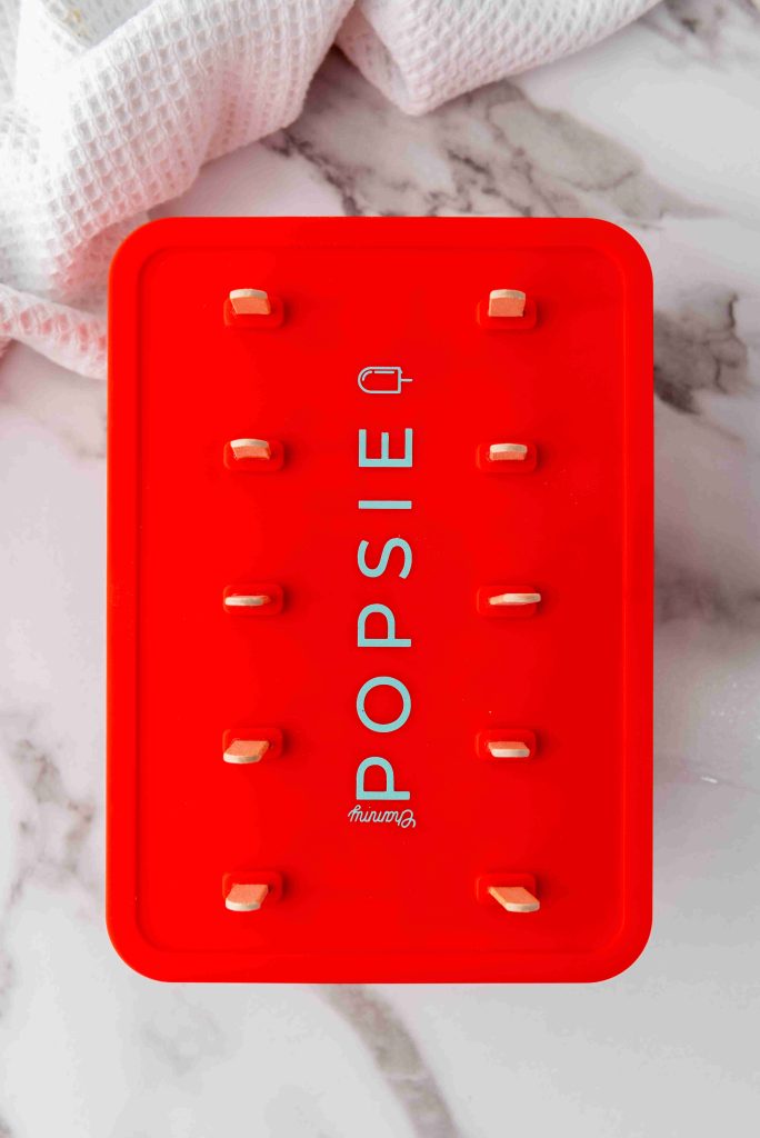 A red popsicle mold labeled POPSIE holds ten wooden sticks, ready for your Strawberry Lemonade Ice Pops, arranged in two rows of five on a white marble surface with a textured white cloth in the corner.