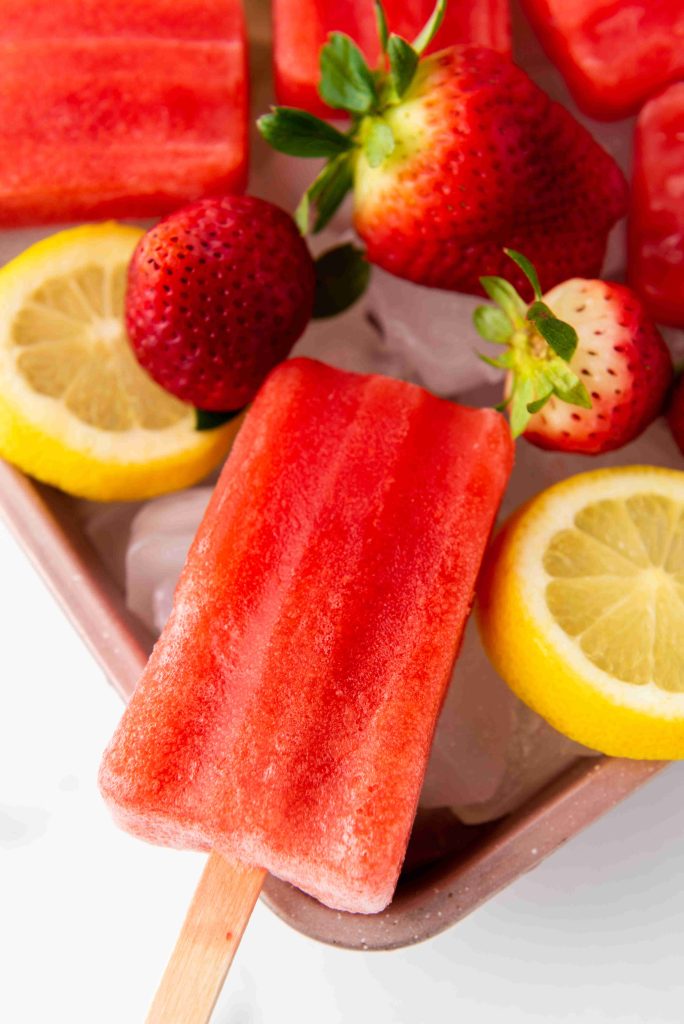 A red Strawberry Lemonade Ice Pop on a stick rests on a tray with ice, fresh strawberries, and lemon slices. The scene is bright and colorful, highlighting this refreshing treat made from an easy strawberry popsicles recipe.