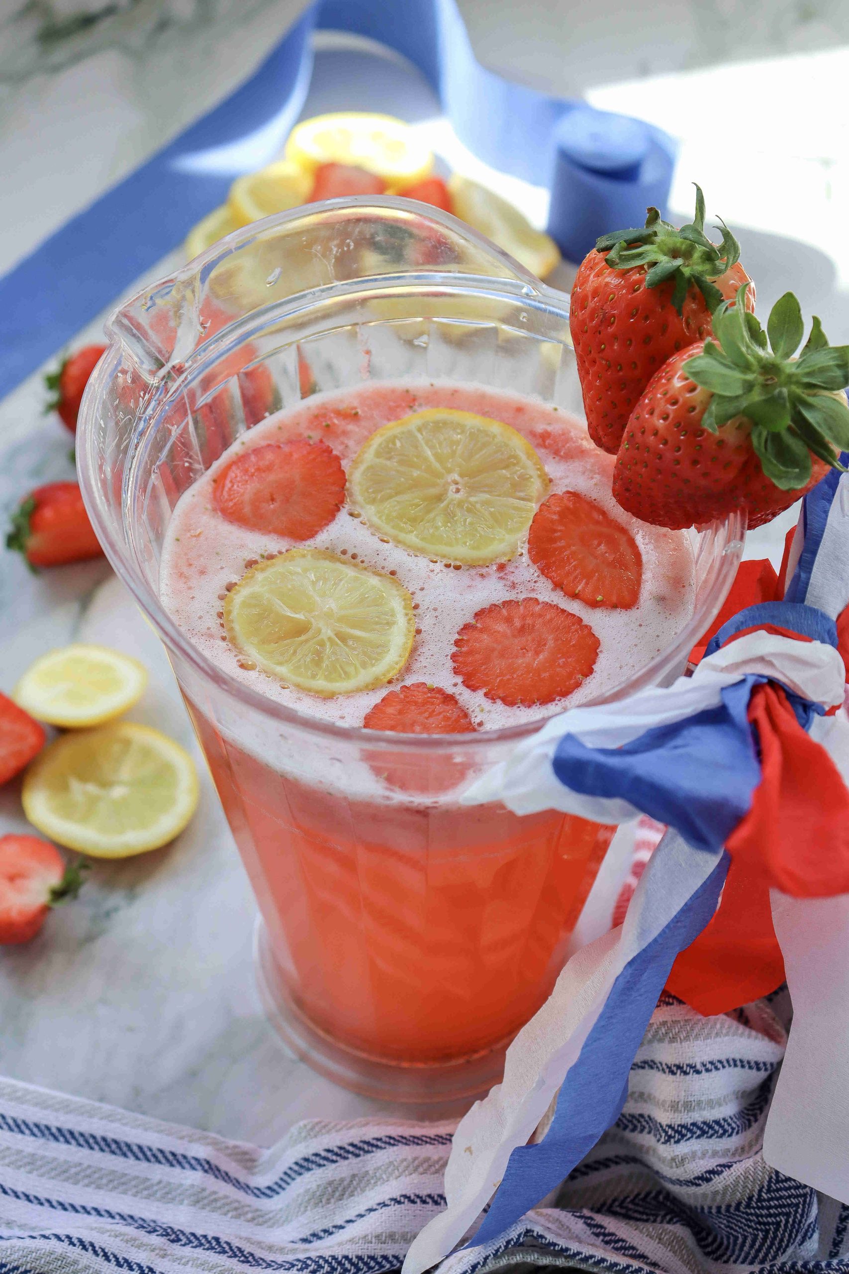 A pitcher of Strawberry Puree Lemonade garnished with lemon and strawberry slices, with two whole strawberries on the rim. Red, white, and blue ribbons are tied around the handle, and sliced lemons and strawberries are nearby.