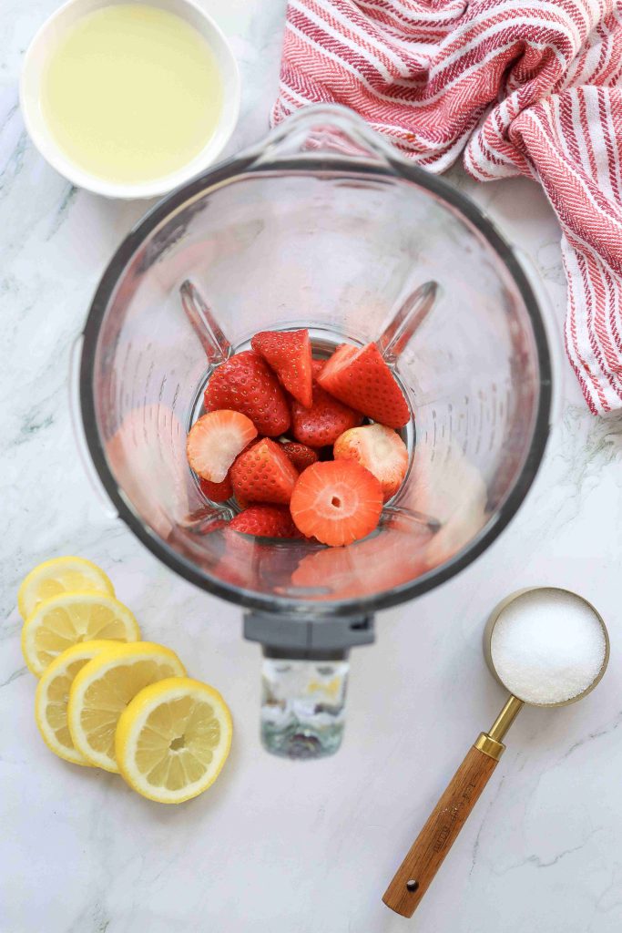 Overhead view of a blender with sliced strawberries inside, surrounded by lemon slices—perfect for making Strawberry Puree Lemonade—along with a bowl of liquid, a measuring cup of sugar, and a red-striped kitchen towel on a marble surface.