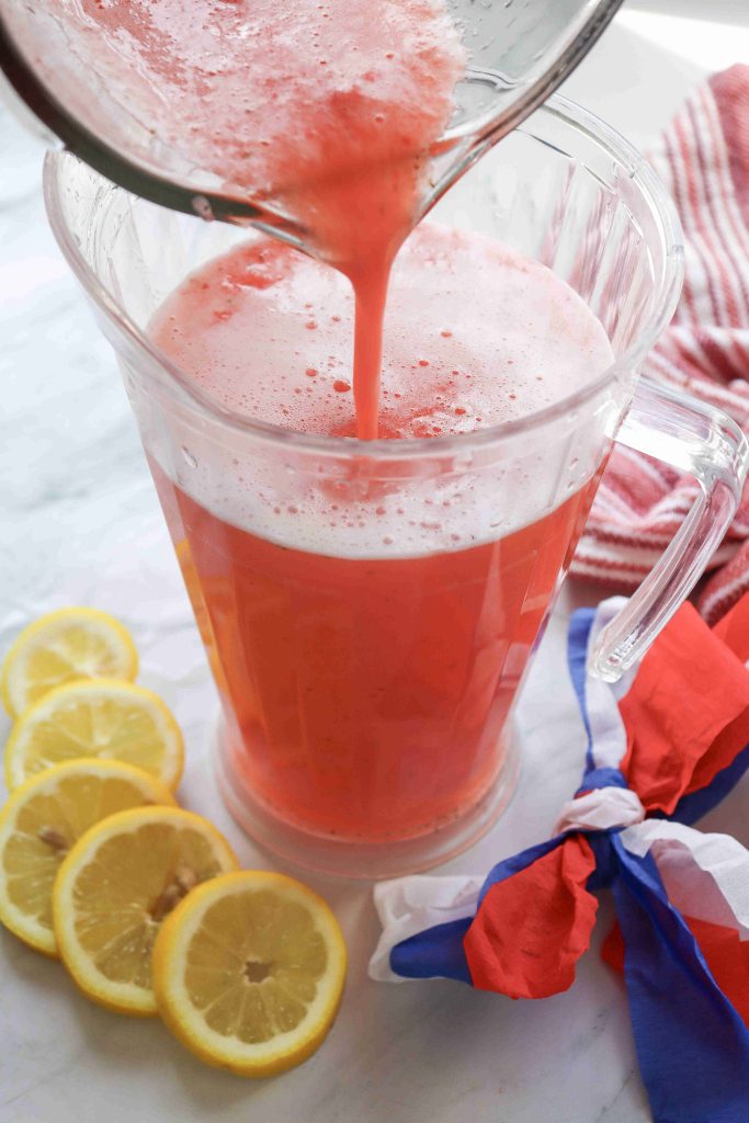 A pitcher is being filled with frothy strawberry puree lemonade, surrounded by lemon slices and a red, white, and blue cloth on a marble surface.