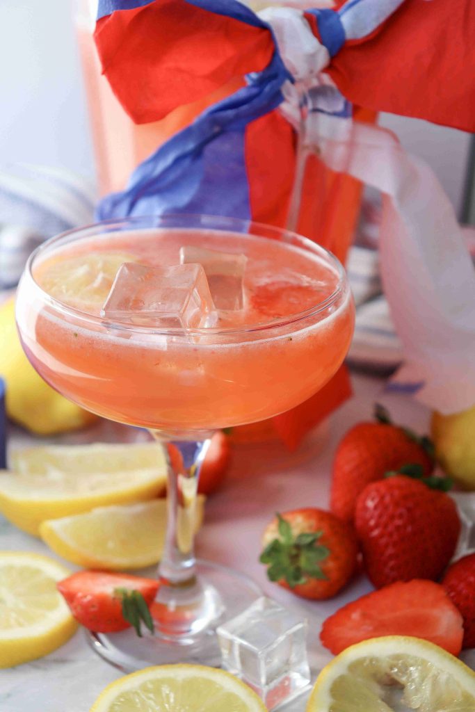 A Strawberry Puree Lemonade cocktail with ice cubes in a coupe glass, surrounded by fresh strawberries, lemon slices, and more ice. In the background, a jar is adorned with festive red, white, and blue ribbons.