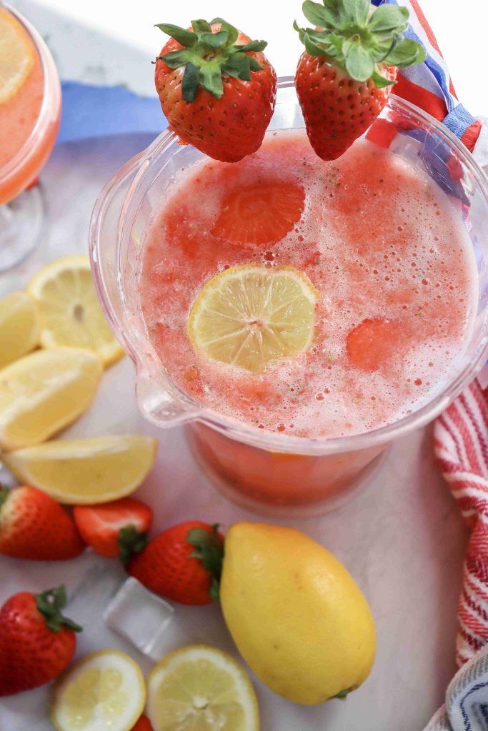 A pitcher of Strawberry Puree Lemonade with lemon and strawberry slices inside, garnished with two whole strawberries on the rim. Surrounded by lemon wedges, whole strawberries, and a striped towel on a white surface.