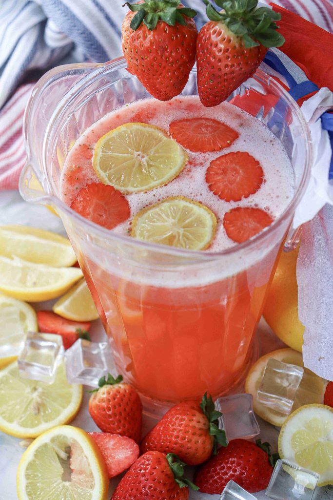 A pitcher of Strawberry Puree Lemonade garnished with lemon and strawberry slices, surrounded by ice cubes, whole strawberries, and lemon slices on a white surface. Two strawberries sit on the rim of the pitcher.