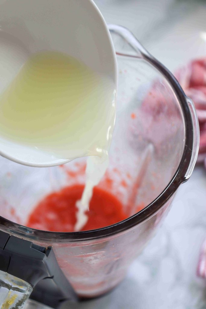 A close-up of a blender with red liquid inside, making Strawberry Puree Lemonade, as a hand pours a white liquid from a small bowl into the blender.